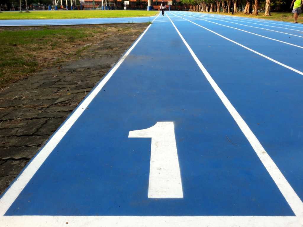 Number 1 / number one lane of running track, white number and white lane lines on blue surface. Low level view with grass runners trees visible in the distance.