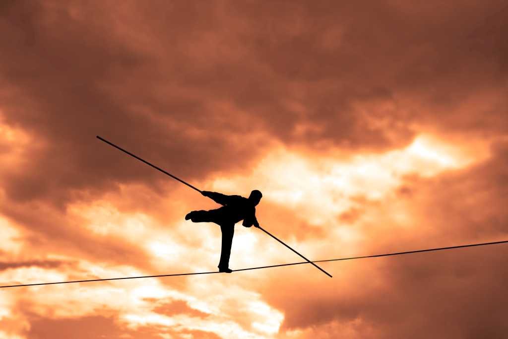 Silhouette of tightrope walker with balancing pole on one leg against a yellow sky background